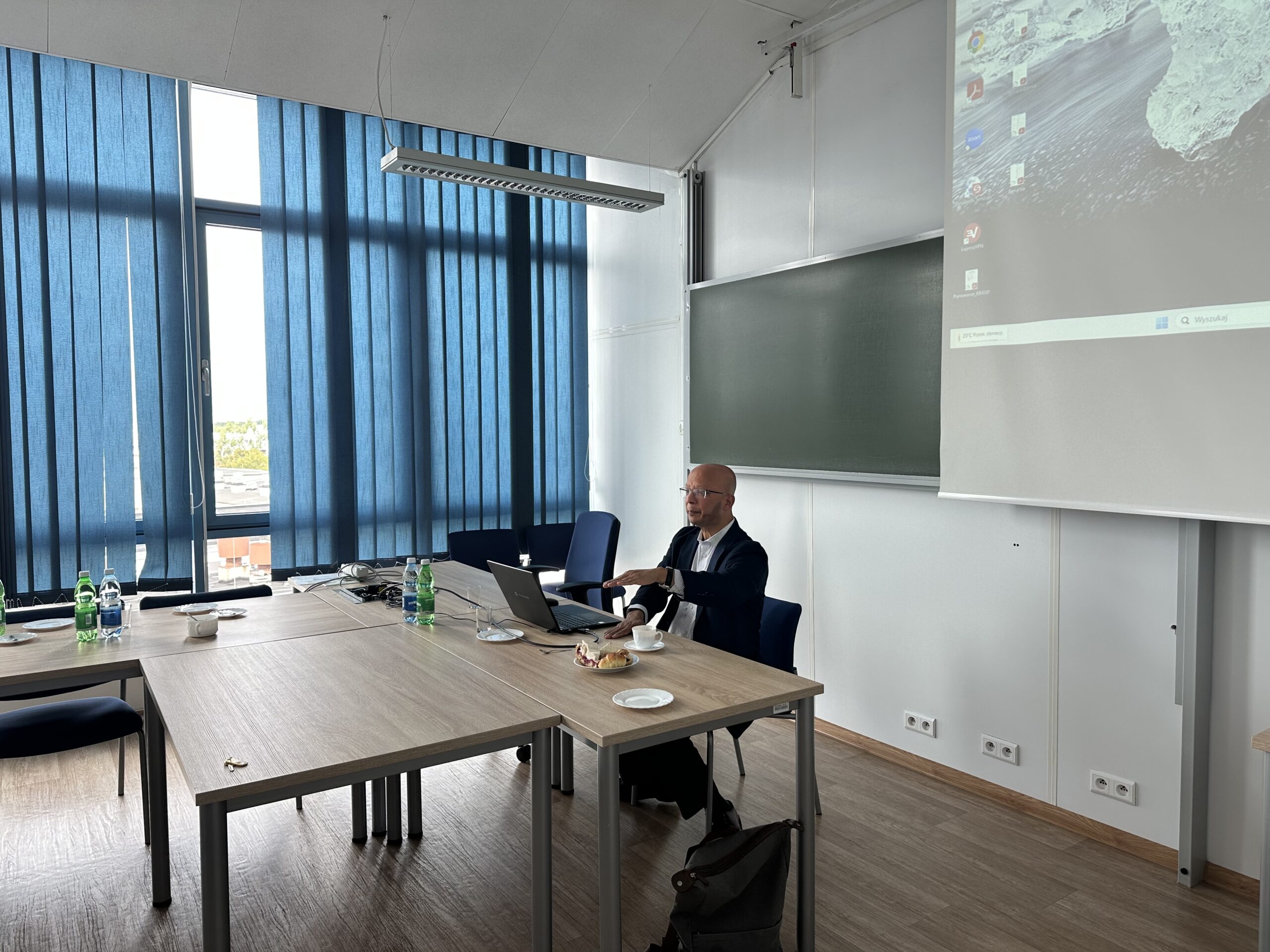 W obrazie przedstawiono nowoczesną salę. Pierwszy plan: - Znajduje się mężczyzna siedzący przy stole, który używa laptopa. - Na stole widoczne są laptopy, kilka butelek z wodą, filiżanki oraz talerzyki z jakimś jedzeniem. - W tle widzimy duże niebieskie żaluzje zasłaniające okna, przez które przechodzi światło dzienne. Na ścianie znajduje się tablica oraz ekran, na którym jest wyświetlany pulpit z projektora. - Jasność obrazu sugeruje, że zdjęcie zostało zrobione w ciągu dnia. Pokój jest przestronny i dobrze oświetlony naturalnym światłem.