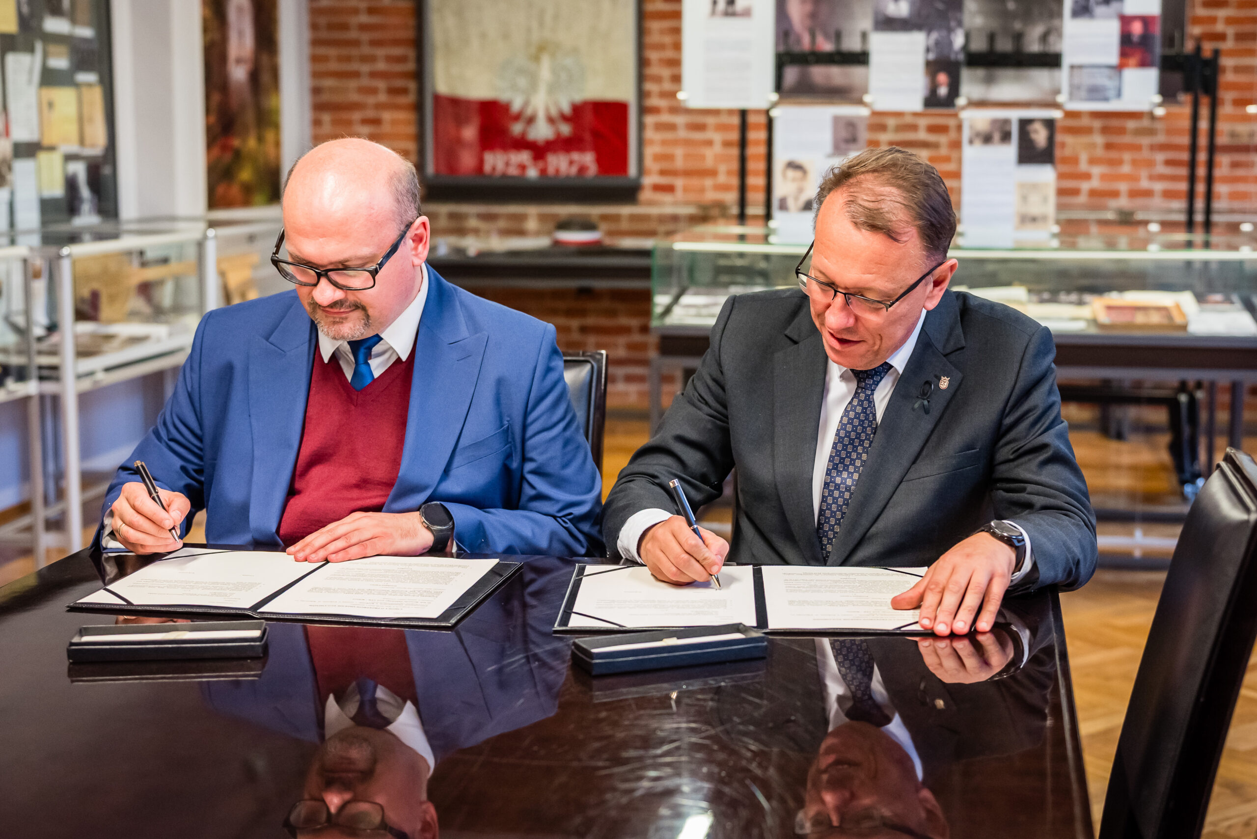 Na pierwszym planie zdjęcia widzimy dwóch mężczyzn siedzących przy ciemnym, błyszczącym stole. Obaj mają przed sobą dokumenty i są zajęci ich podpisywaniem. Mężczyzna po lewej stronie nosi niebieską marynarkę, bordowy sweter i niebieski krawat. Ten po prawej stronie ma na sobie ciemny garnitur, białą koszulę i wzorzysty krawat. Obaj skoncentrowani są na czynności, którą wykonują. W tle znajdują się gabloty z dokumentami oraz tablice informacyjne, co sugeruje, że zdjęcie wykonano w miejscu związanym z historią lub edukacją, takim jak muzeum czy archiwum. Na jednej ze ścian wisi flaga z orłem, co może wskazywać na polskie instytucje. Atmosfera zdjęcia jest poważna i formalna, podkreślona przez elegancki ubiór mężczyzn i charakter wnętrza. Oświetlenie jest jasne, a zdjęcie wygląda na wykonane w ciągu dnia.
