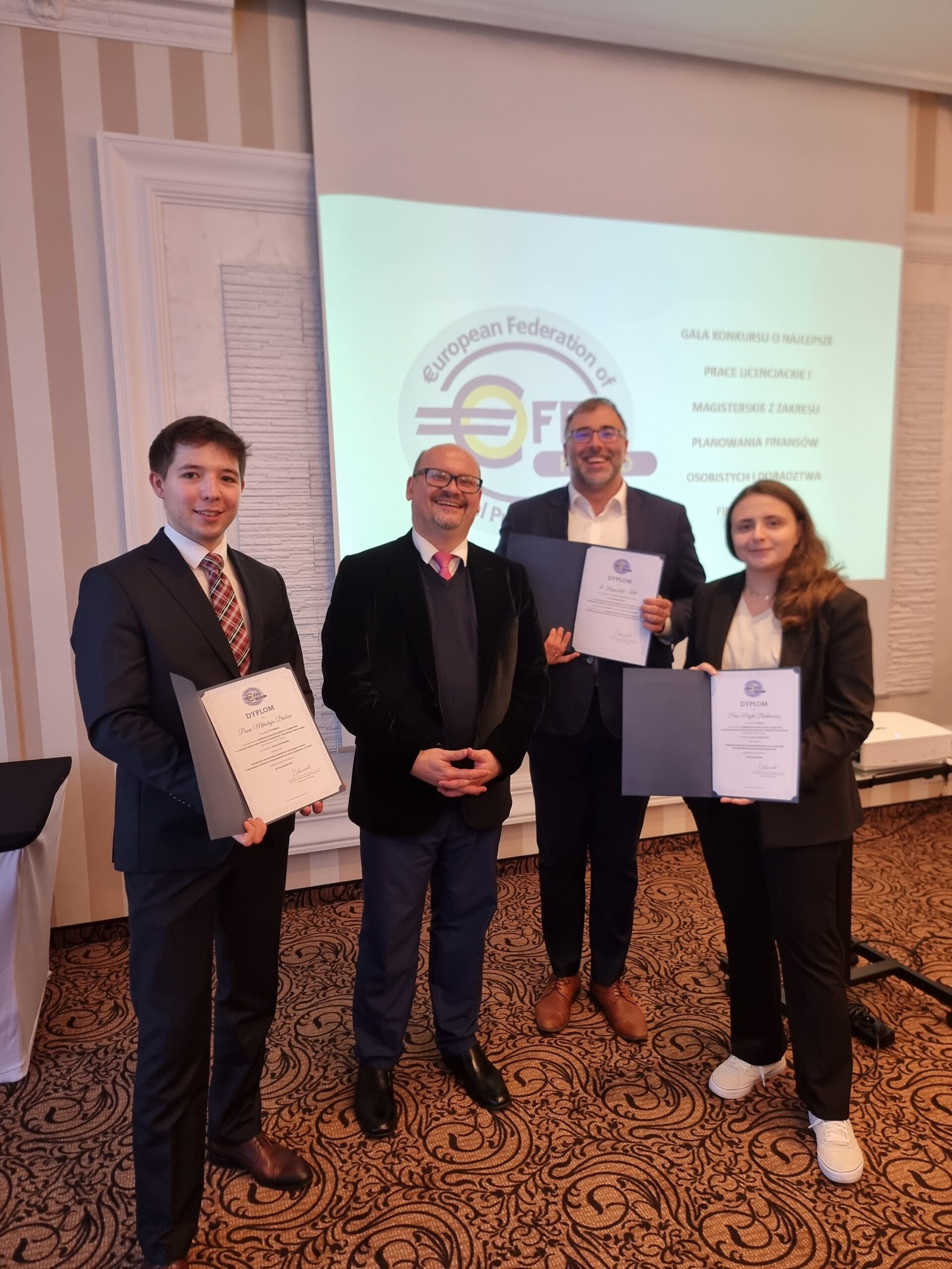 Na pierwszym planie zdjęcia widocznych jest czterech osób, trzy z nich trzymają dyplomy. Osoby te są elegancko ubrane. W tle widać ekran z logo i napisami. Sceneria wskazuje, że jest to wnętrze sali konferencyjnej. Atmosfera zdjęcia jest uroczysta i oficjalna, co podkreślone jest formalnym strojem obecnych osób i dyplomami, które trzymają. Stylizacje wnętrza oraz sposób ubioru wskazują na formalne wydarzenie. Nie ma widocznych oznak, które mogłyby dokładnie określić porę dnia czy roku, ale obecność światła sugeruje, że zdjęcie zostało zrobione w ciągu dnia.