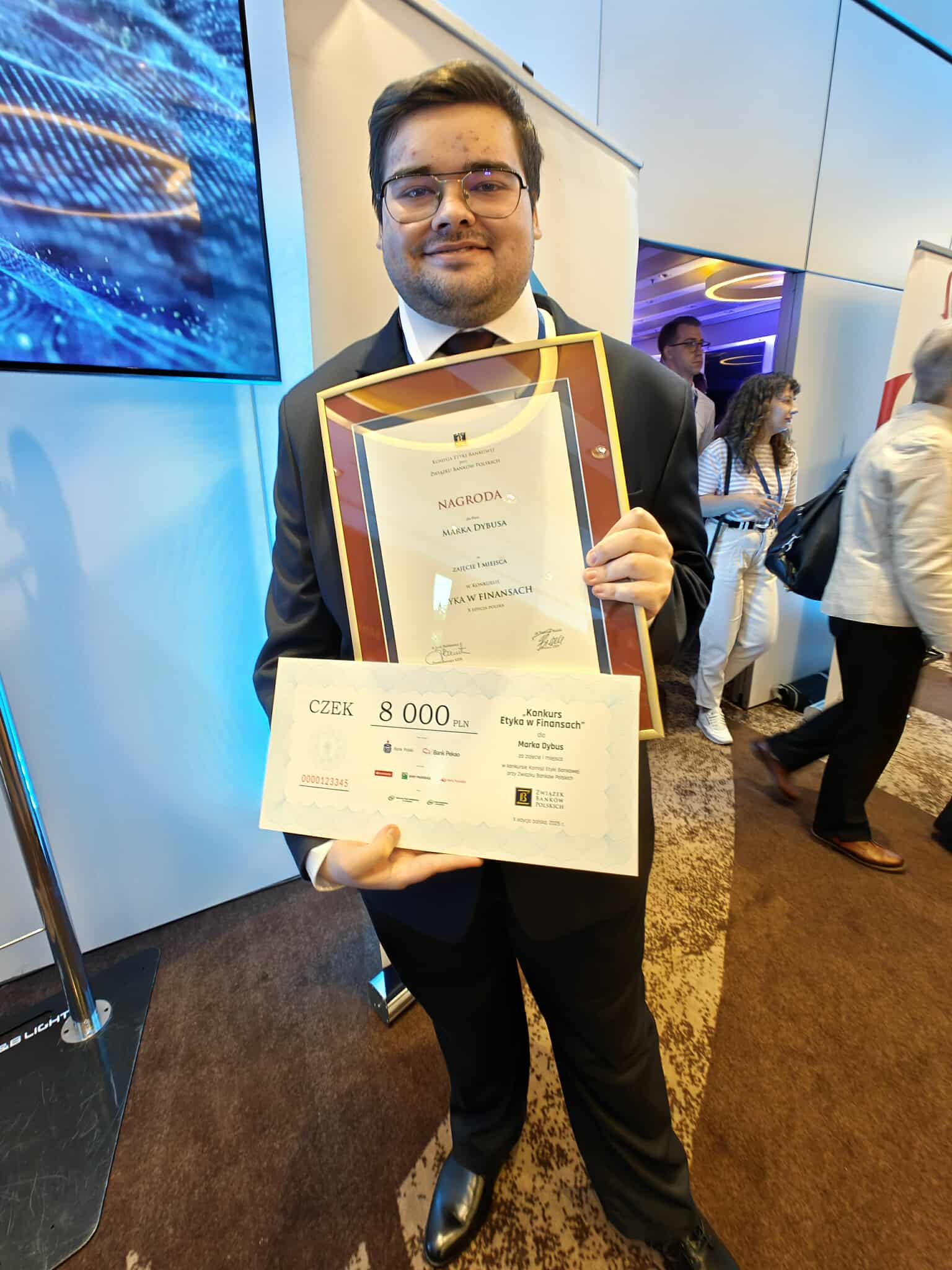 Zdjęcie przedstawia osobę w garniturze trzymającą dyplom oraz czek. W tle po lewej stronie widać telewizor. Z kolei po prawej stronie widać ludzi wchodzących do sali.