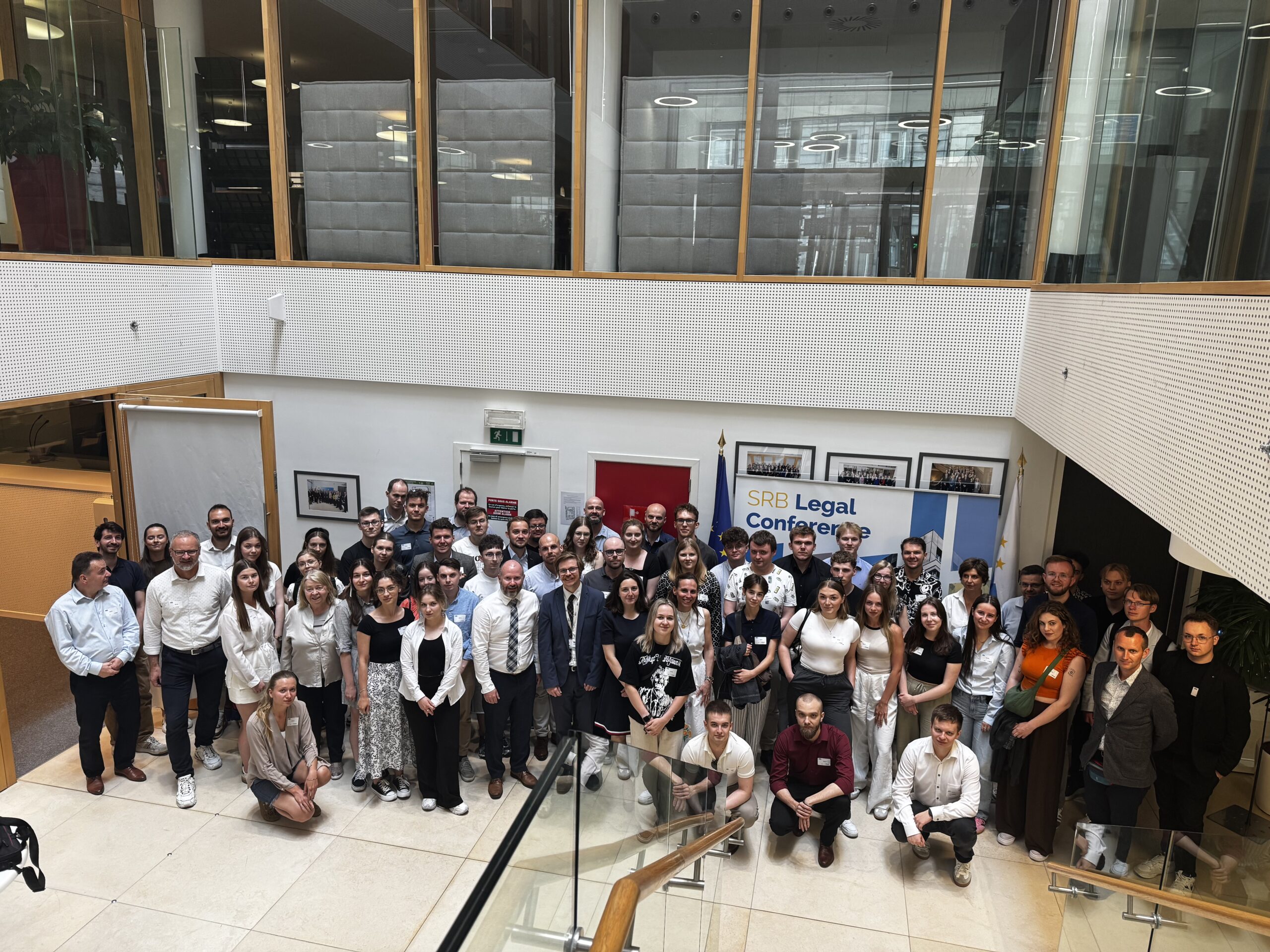Na zdjęciu widoczna jest duża grupa osób stojąca we wnętrzu nowoczesnego budynku, pozująca do wspólnego zdjęcia. To uczestnicy wyjazdu studyjnego do Brukseli wraz z ekspertami ds. restrukturyzacji i uporządkowanej likwidacji banku z Single Resolution Board. Większość osób uśmiecha się i patrzy w stronę obiektywu. Ubrania są głównie formalne i półformalne. W tle znajdują się przeszklone i drewniane elementy architektoniczne, a także banner z napisem "SRB Legal Conference", podkreślający oficjalny charakter wydarzenia.