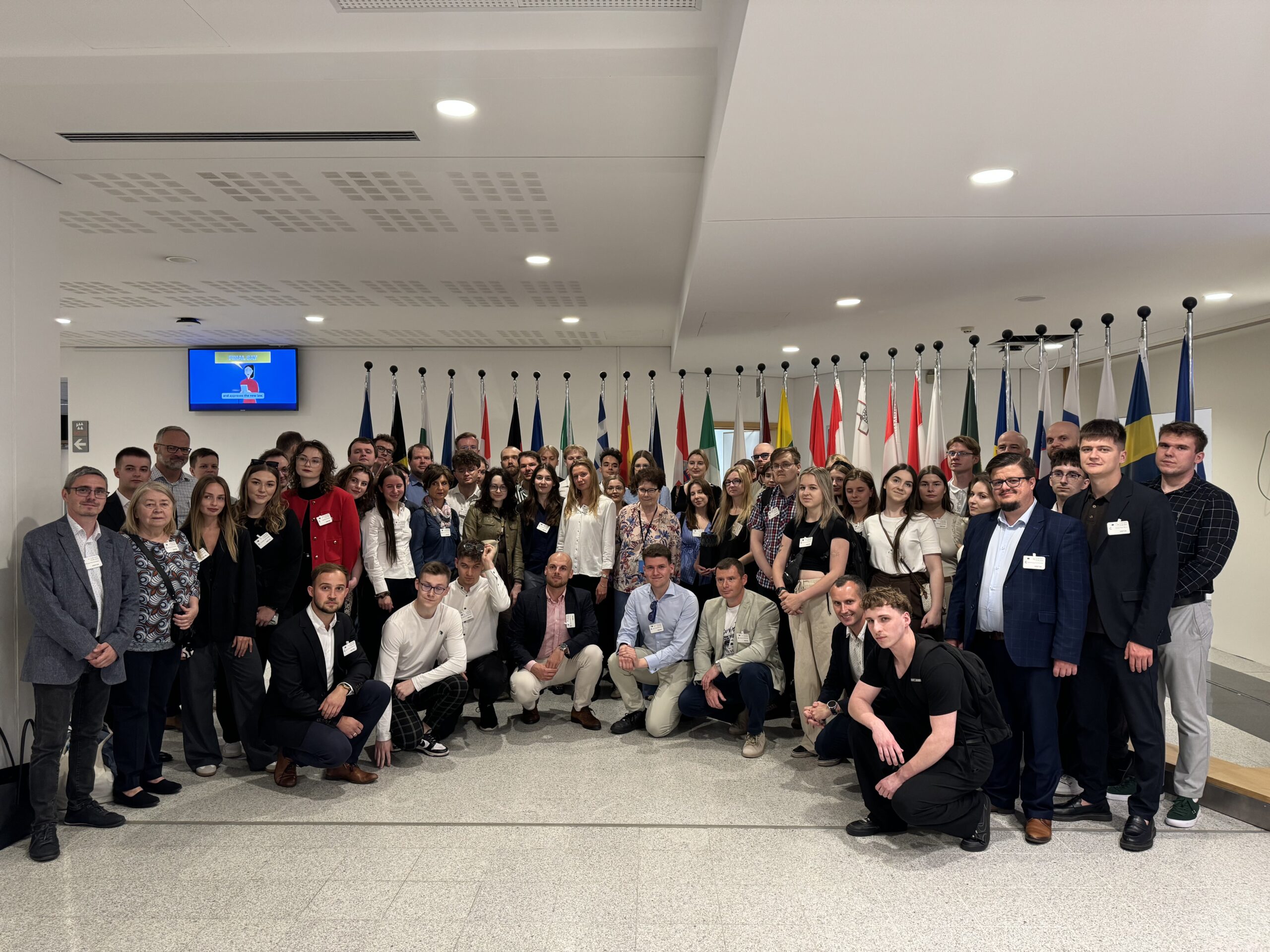Na zdjęciu znajduje się duża grupa osób pozujących do wspólnego zdjęcia w dobrze oświetlonym pomieszczeniu. Są to uczestnicy wyjazdu studyjnego do Brukseli wraz z Panią Europoseł Hanną Gronkiewicz-Waltz, a zdjęcie zostało wykonane w Parlamencie Europejskim. W tle widoczne są flagi różnych państw członkowskich UE oraz monitor wyświetlający obraz, co podkreśla oficjalny charakter miejsca. Uczestnicy ubrani są zarówno w stroje formalne, jak i bardziej casualowe.