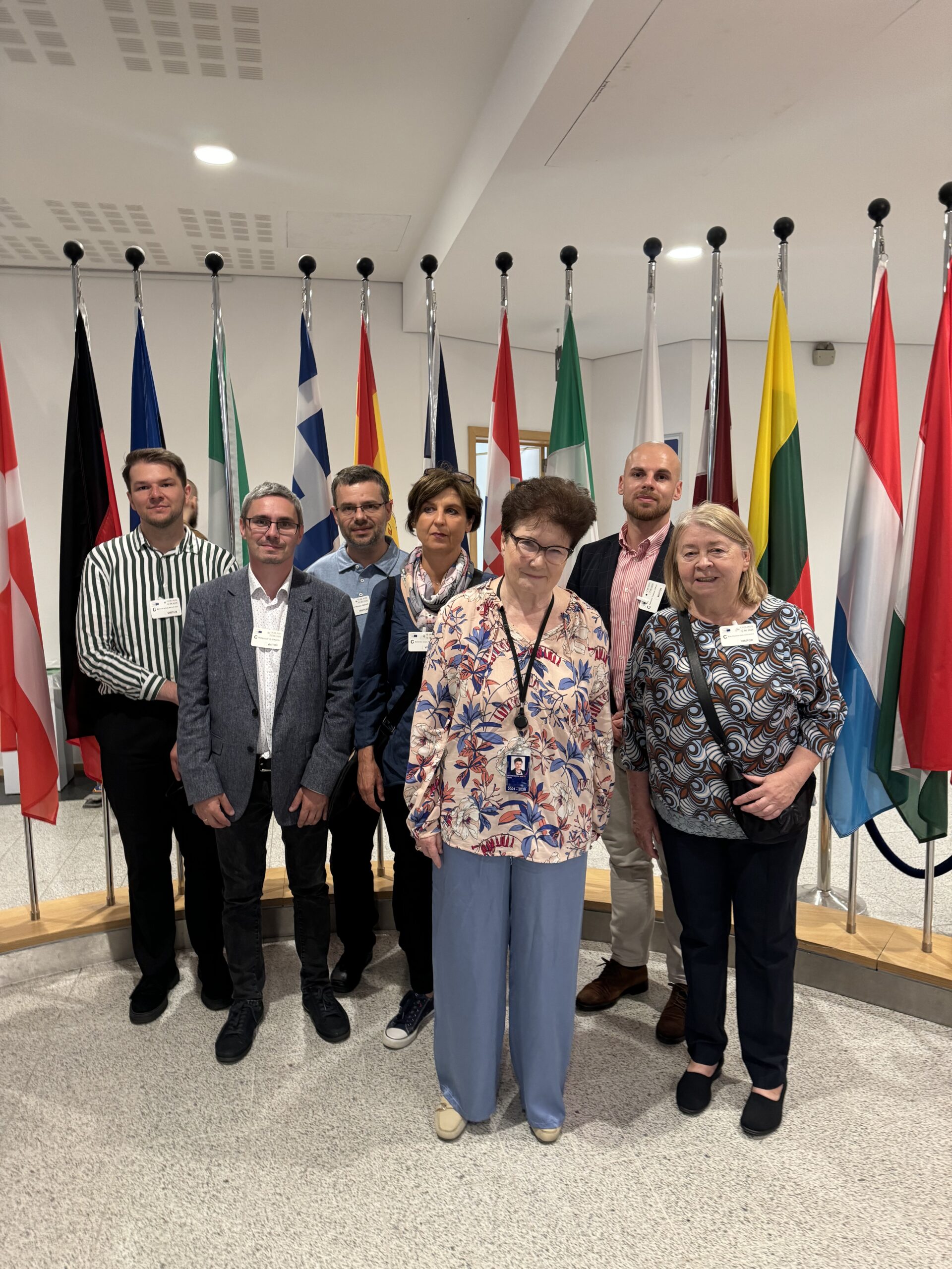 Na zdjęciu znajduje się grupa siedmiu osób, pozujących do zdjęcia grupowego na tle rzędu flag narodowych, wskazujących na instytucję związaną z Unią Europejską. Wśród nich są pracownicy KBiGSF oraz Pani Prof. Hanna Gronkiewicz-Waltz – była Prezes Narodowego Banku Polskiego, wiceprezes Europejskiego Banku Odbudowy i Rozwoju, prezydent Warszawy, a obecnie członkini Komisji Gospodarczej i Monetarnej (ECON) Parlamentu Europejskiego.