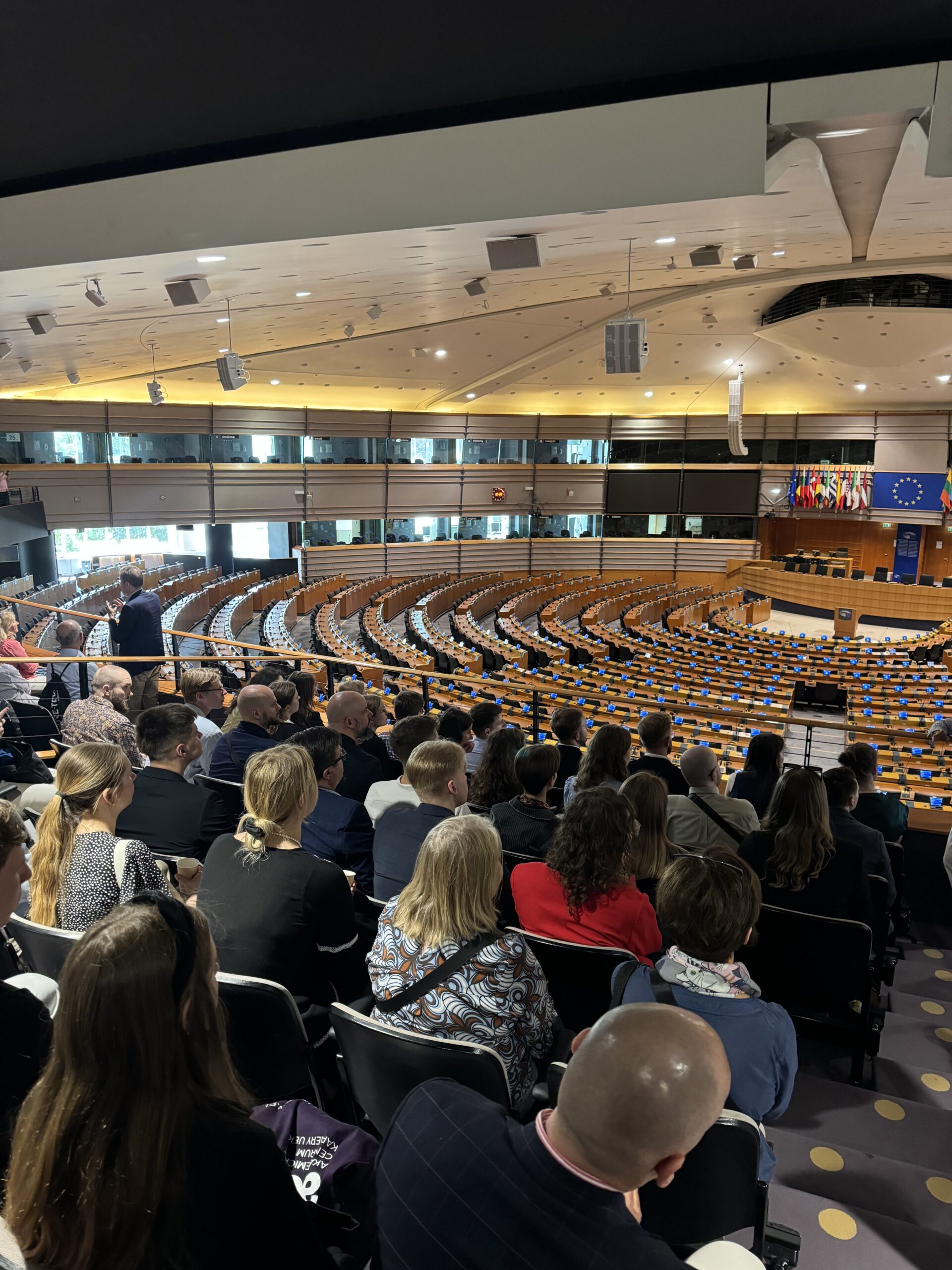 Widok z galerii dla publiczności na salę obrad Parlamentu Europejskiego w Brukseli. Na pierwszym planie siedzi grupa osób, uczestników wyjazdu, obserwujących wnętrze sali. Przed nimi znajdują się rzędy pustych miejsc dla europosłów, każdy z mikrofonem i tabliczką. W tle widać podium mówcy, flagi państw Unii Europejskiej oraz flagę UE z 12 gwiazdami. Całość znajduje się w nowoczesnym, przestronnym wnętrzu o oświetleniu wpuszczanym przez okna i sztucznym świetle sufitowym.